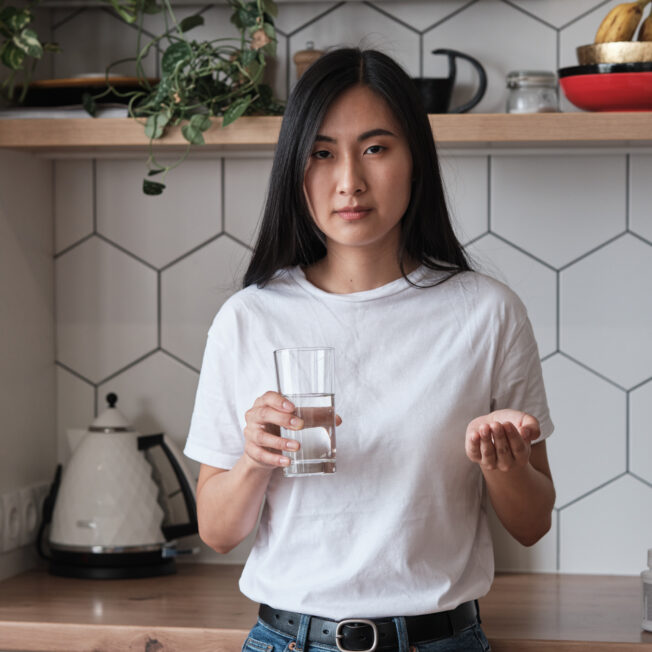 an Asian person with long hair holds a glass of water and a pill.