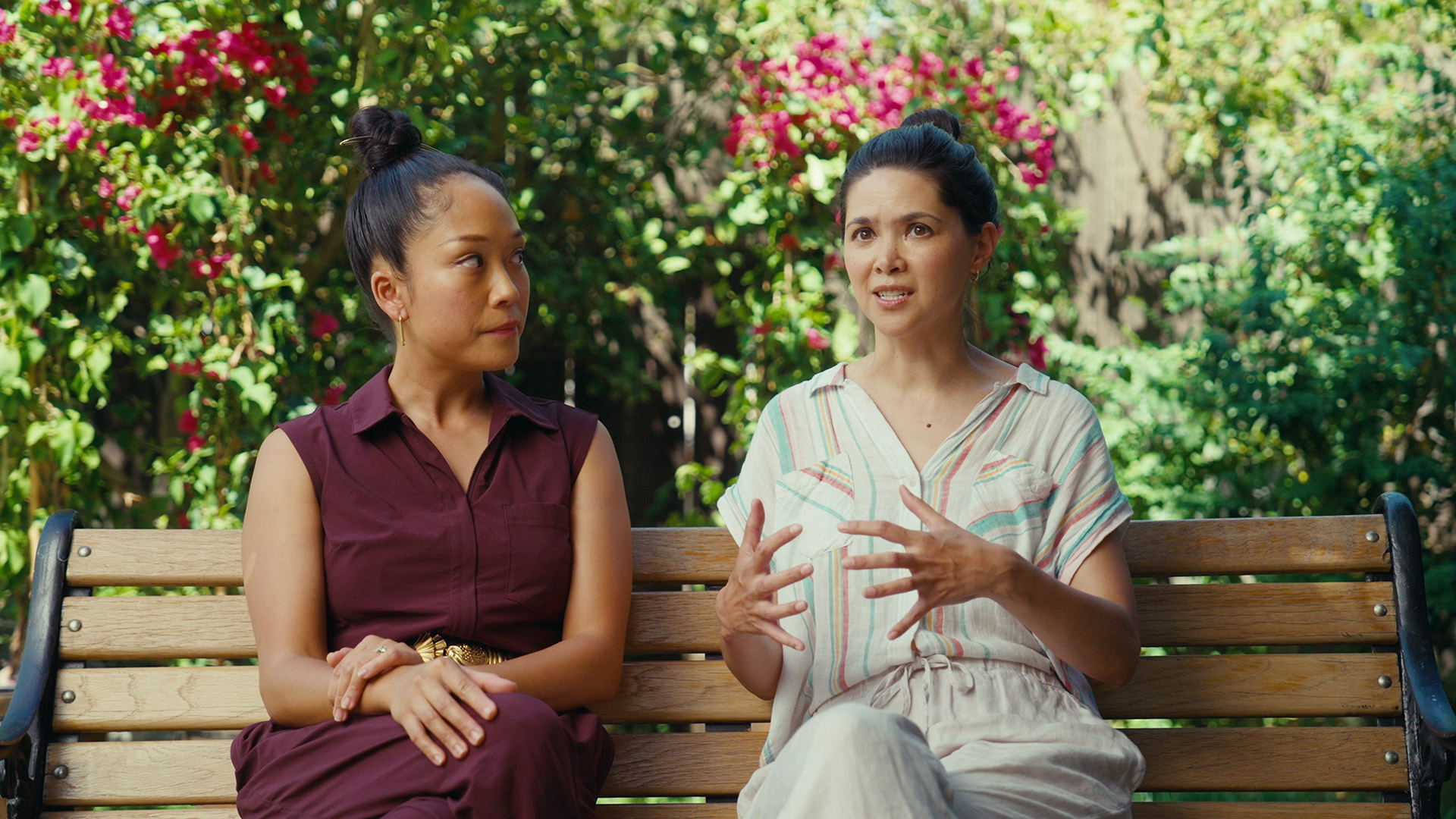 Two AAPI women sit on a bench in an outdoor park while chatting.