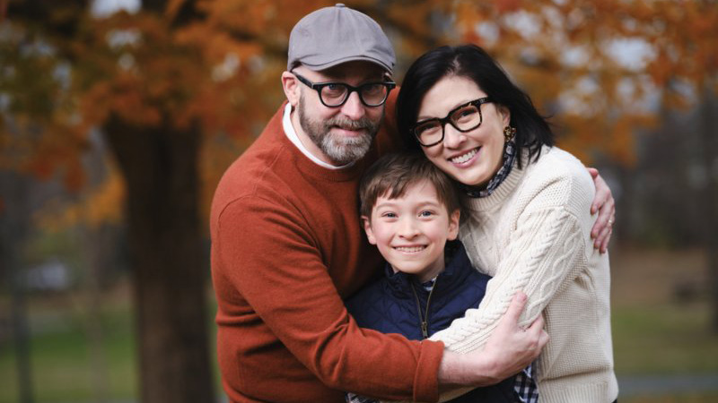 Virginia Yuna, her husband, and her son embrace outdoors with autumn foliage in the background.