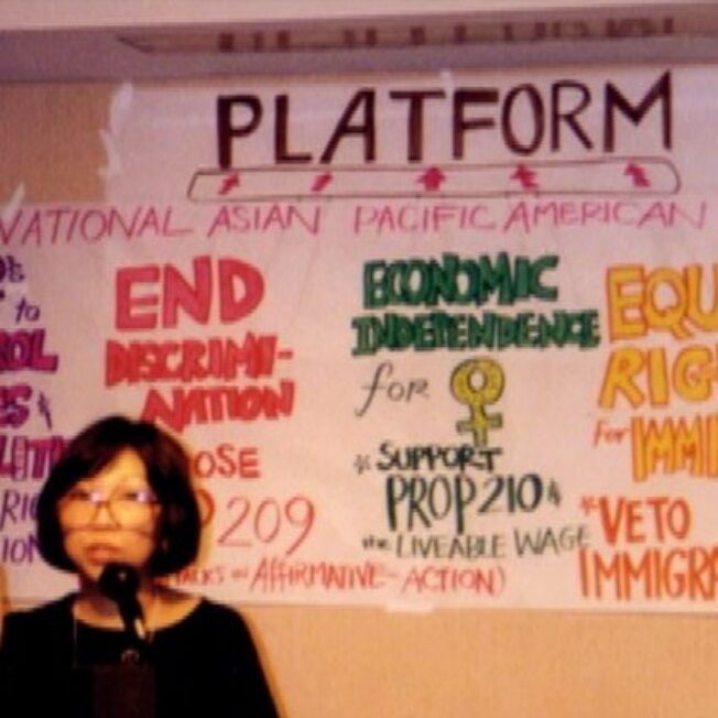 A woman speaks at a podium. A hand-written poster in the background diagrams the early NAPAWF policy platform, including reproductive rights, antidiscrimination, economic independence, immigrant rights, and ending violence against women.